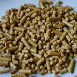 Jasne pellety drzewne ułożone w stos na białym tle, widoczne drobne wióry i nierówności w strukturze pelletu.