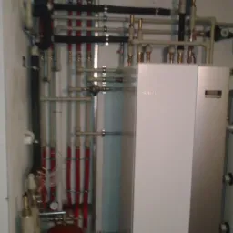 Złożona instalacja grzewcza z rurami w kolorze czerwonym i szarym, prowadząca do białego pieca grzewczego w pomieszczeniu technicznym.