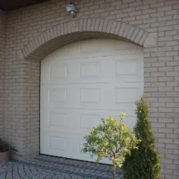 Biała segmentowa brama garażowa z ceglanym łukiem nad nią, otoczona niskimi krzewami i kostką brukową.