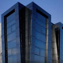 Nowoczesna fasada budynku biurowego z ciemnego szkła i metalu, odbijająca niebo, widziana z dołu pod kątem, ukazująca geometryczny design i modularną konstrukcję.