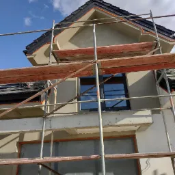 Dom w trakcie prac wykończeniowych elewacji, widoczne rusztowania, zabezpieczone okna folią i świeżo nałożony tynk strukturalny na szczycie budynku.