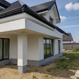 Nowoczesny, dwukondygnacyjny dom z jasną elewacją i ciemną dachówką, widok z boku, z widocznym fragmentem ogrodu i sąsiedniego budynku w tle.