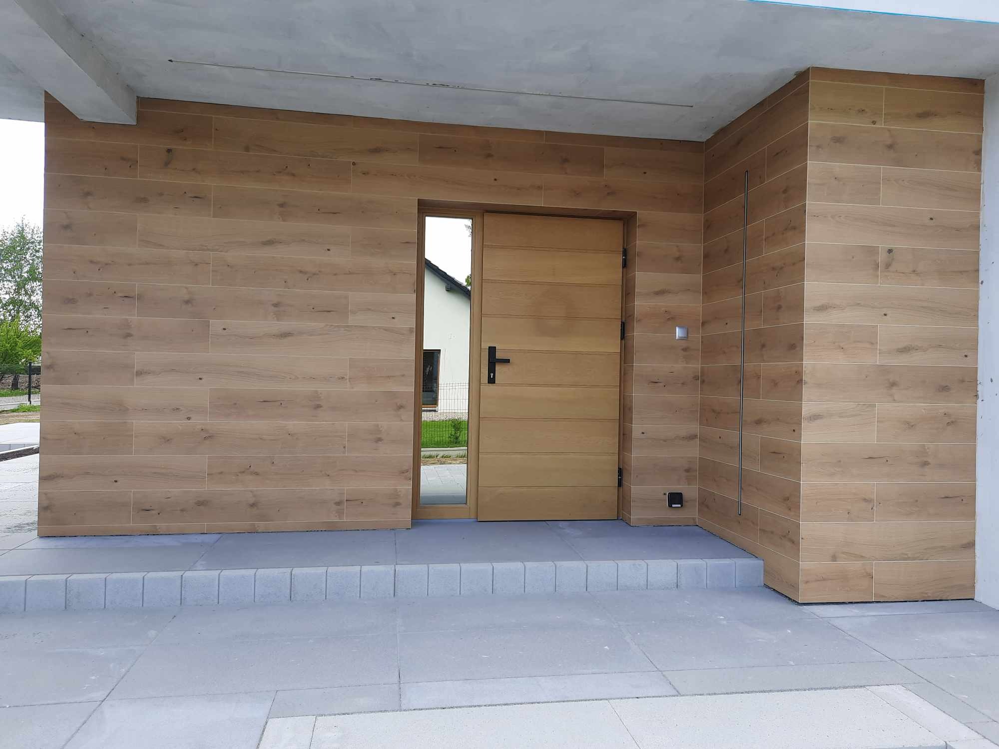 Wejście do domu wykończone płytkami imitującymi drewno, z jasnymi, drewnianymi drzwiami z czarną klamką i szarą kostką brukową na podjeździe.