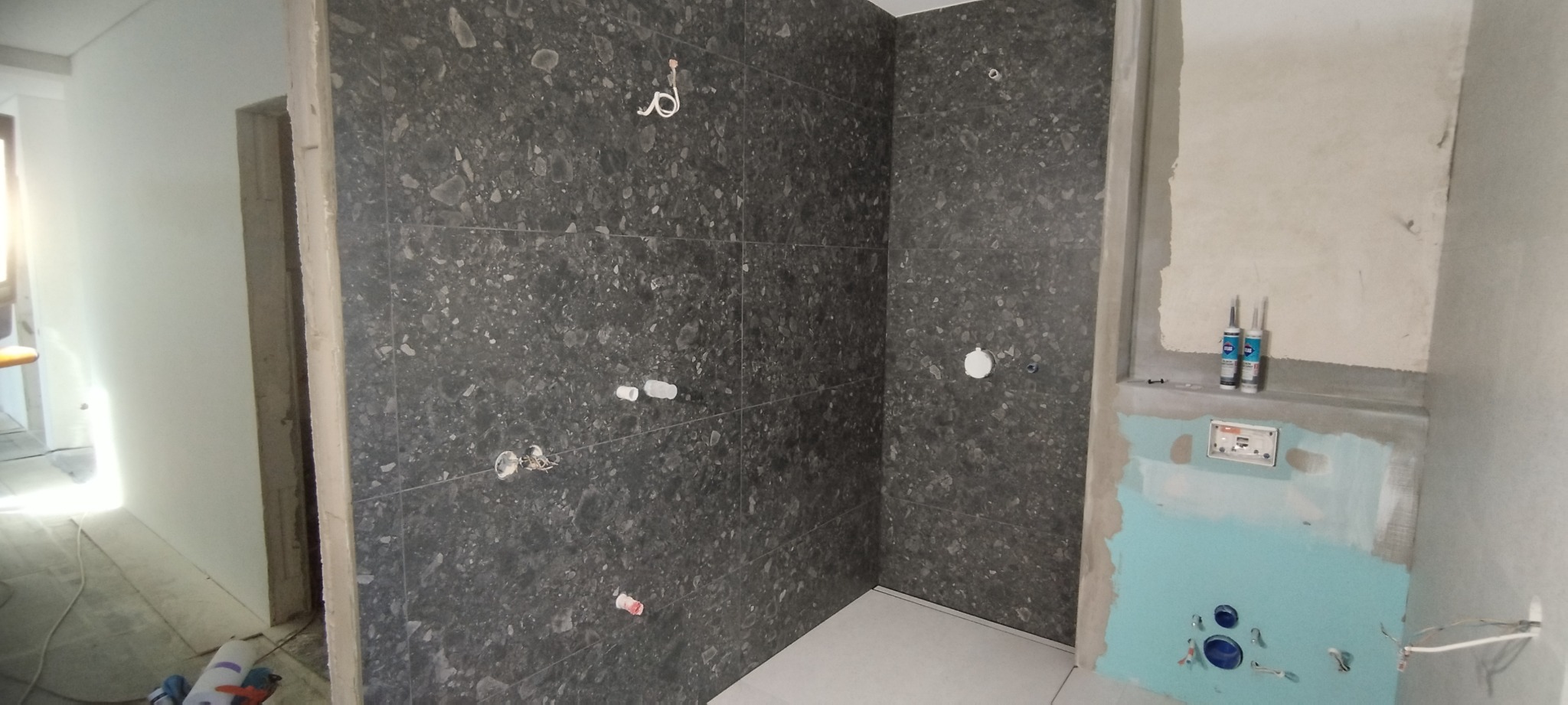 Wnętrze łazienki w trakcie remontu, z ciemnymi płytkami imitującymi kamień na ścianach, widoczne przygotowane punkty instalacyjne, puszka podtynkowa, oraz dwie tuby silikonu na półce...
