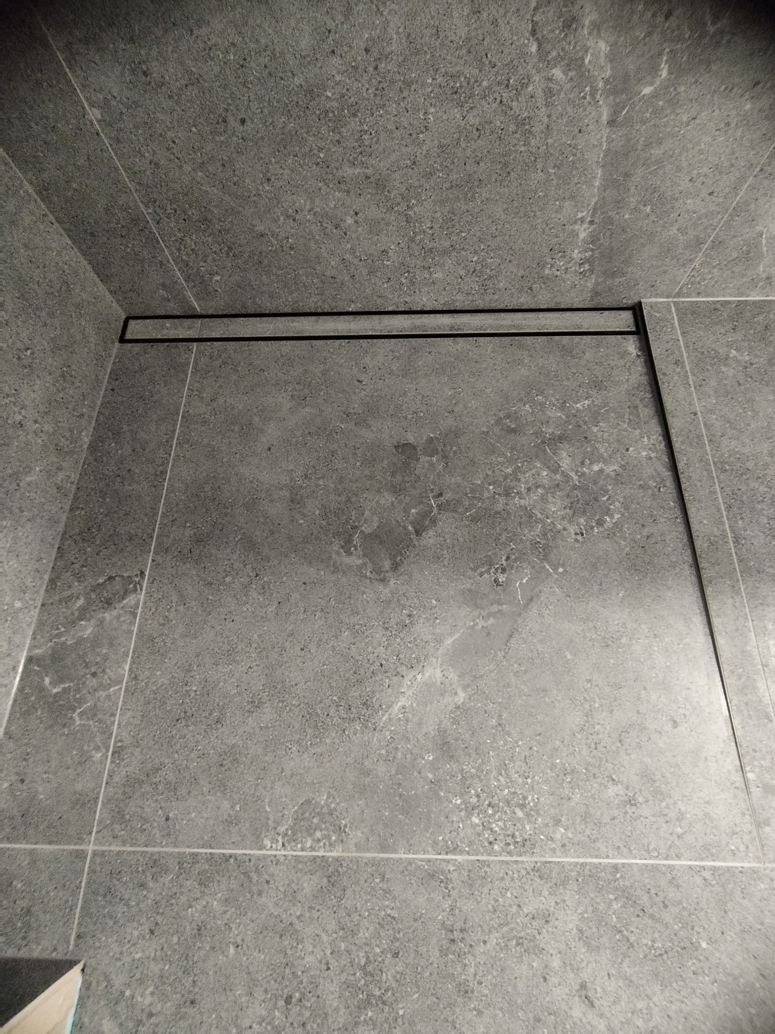 Szare płytki podłogowe i ścienne w kabinie prysznicowej z czarnym odpływem liniowym, widok z góry.