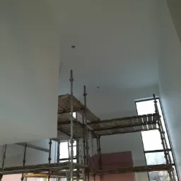 Wnętrze w Jarocinie w trakcie malowania, widoczne rusztowanie i świeżo pomalowane ściany na biało, z widocznymi otworami na oświetlenie.