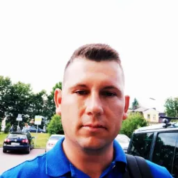 Portret mężczyzny w niebieskiej koszulce polo na tle miejskiego krajobrazu z samochodami i zielenią. Ujęcie z dołu, oświetlenie dzienne.