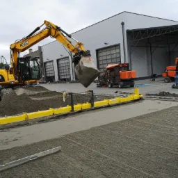 Żółta koparka JCB z pełną łyżką ziemi nad przygotowanym podłożem pod kostkę, widoczne maszyny do układania kostki, wyrównane podłoże i ułożone obrzeża.