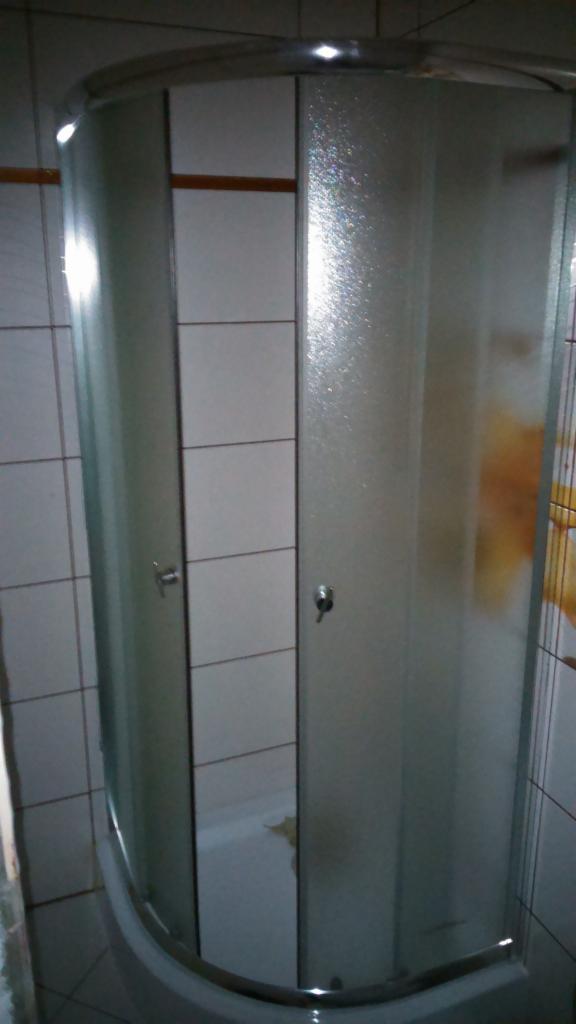 Kabina prysznicowa z przesuwnymi, matowymi drzwiami, widoczna na tle białych płytek z ciemnymi fugami. Chromowane wykończenia i uchwyty.