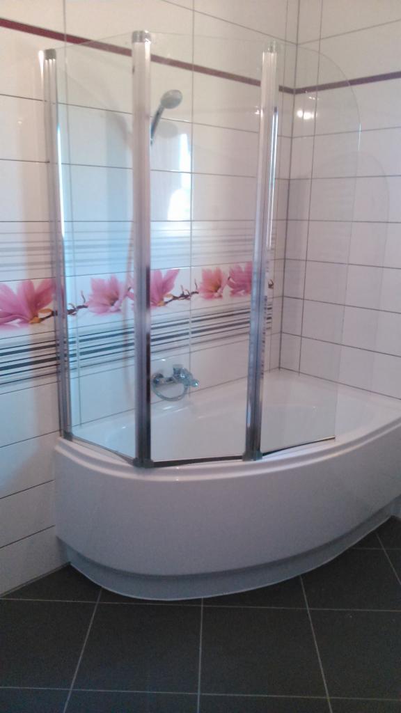 Biała wanna z zamontowanym, półokrągłym parawanem nawannowym z przezroczystego szkła. Na ścianie białe płytki z poziomym paskiem z motywem kwiatowym.