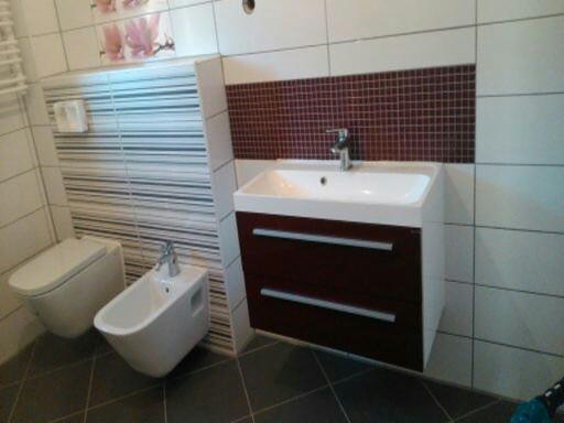 Nowoczesna łazienka po remoncie: biała ceramika (toaleta, bidet) i umywalka z szafką w odcieniach brązu na tle płytek w różnych wzorach.