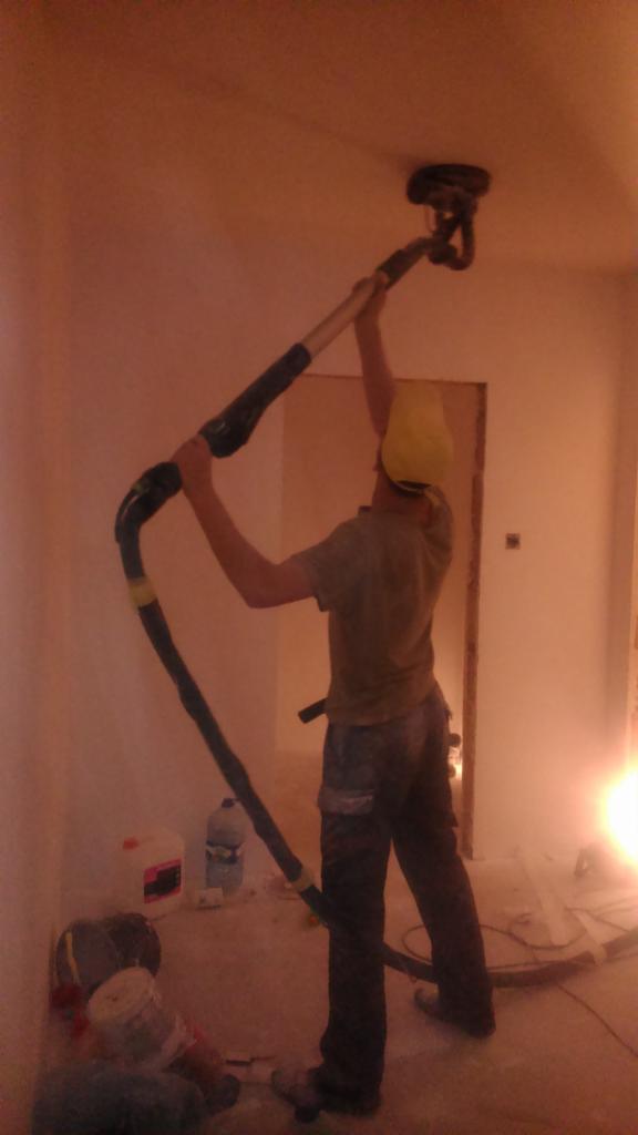 Pracownik w żółtej czapce szlifuje sufit za pomocą szlifierki z odkurzaczem. Widoczne ślady szlifowania. W tle butelka wody i pojemnik z farbą.