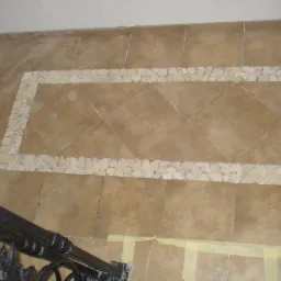 Beżowa podłoga wyłożona kwadratowymi płytkami z dekoracyjnym obramowaniem z jasnych, nieregularnych kamieni, widoczne fragmenty czarnej, metalowej balustrady schodów i żółta taśma malarska.