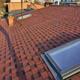 Czerwono-czarny dach pokryty gontem bitumicznym z dwoma oknami dachowymi, widok z góry, w tle budynki mieszkalne.