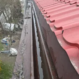 Czerwony dach z systemem kolców przeciw ptakom na rynnie, która jest wypełniona wodą i zanieczyszczeniami, widok z góry na otoczenie z elementami rusztowania.