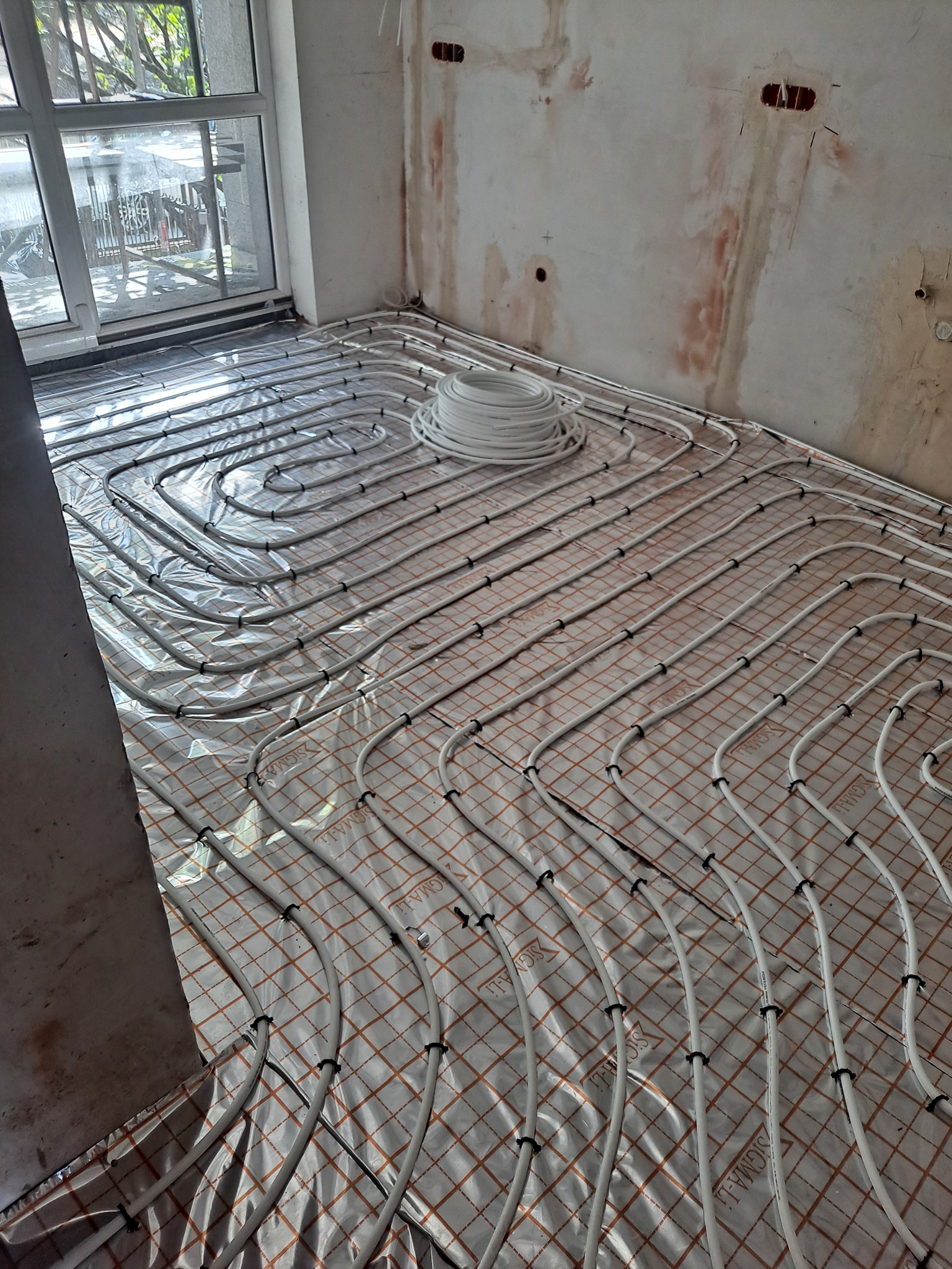 Rozłożone rury ogrzewania podłogowego na izolacji w surowym pomieszczeniu przed wylewką, widoczny zwój rur i okno.