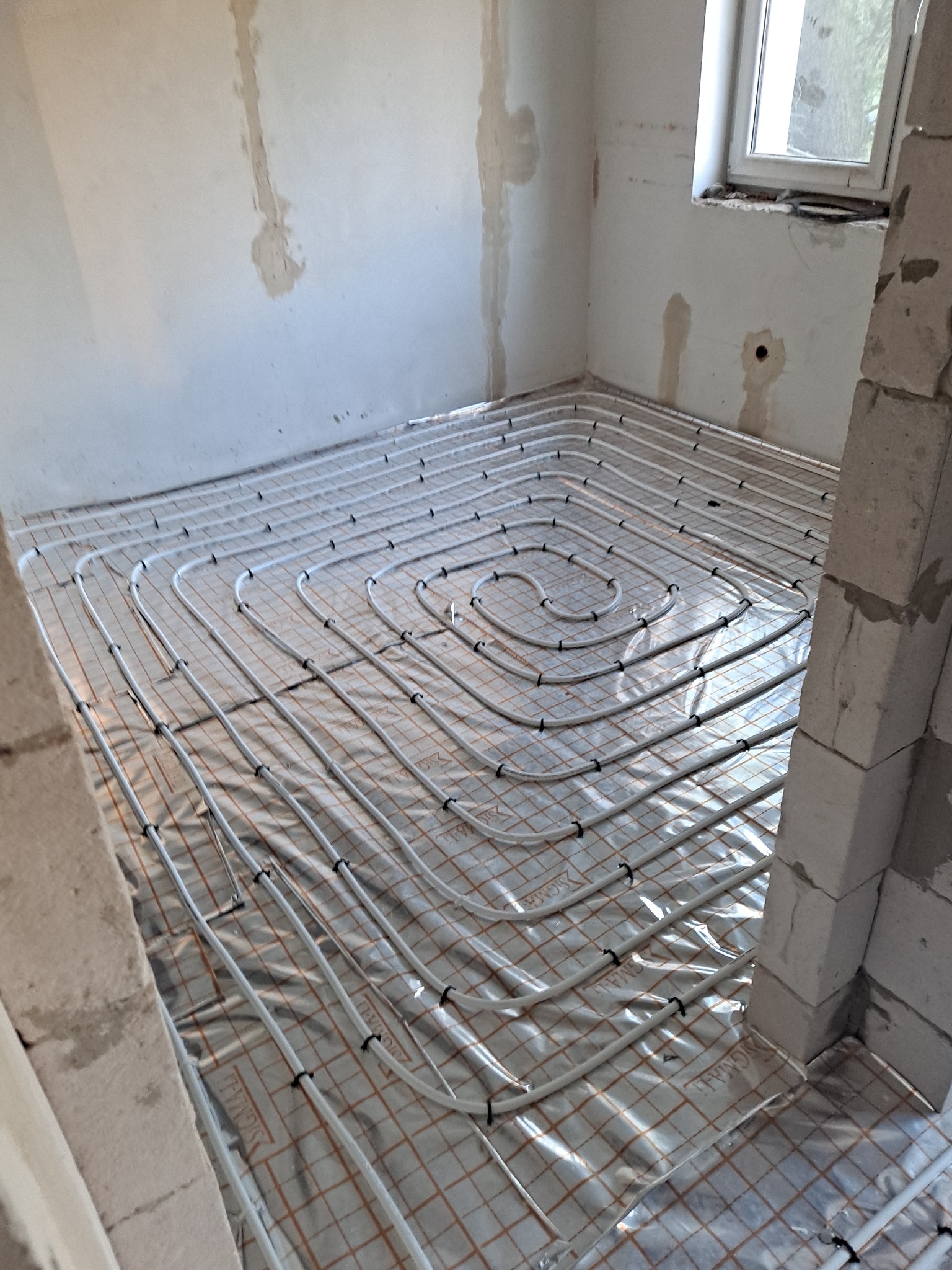 Białe rurki ogrzewania podłogowego ułożone spiralnie na folii z siatką w surowym wnętrzu z oknem i fragmentem ściany z pustaków.