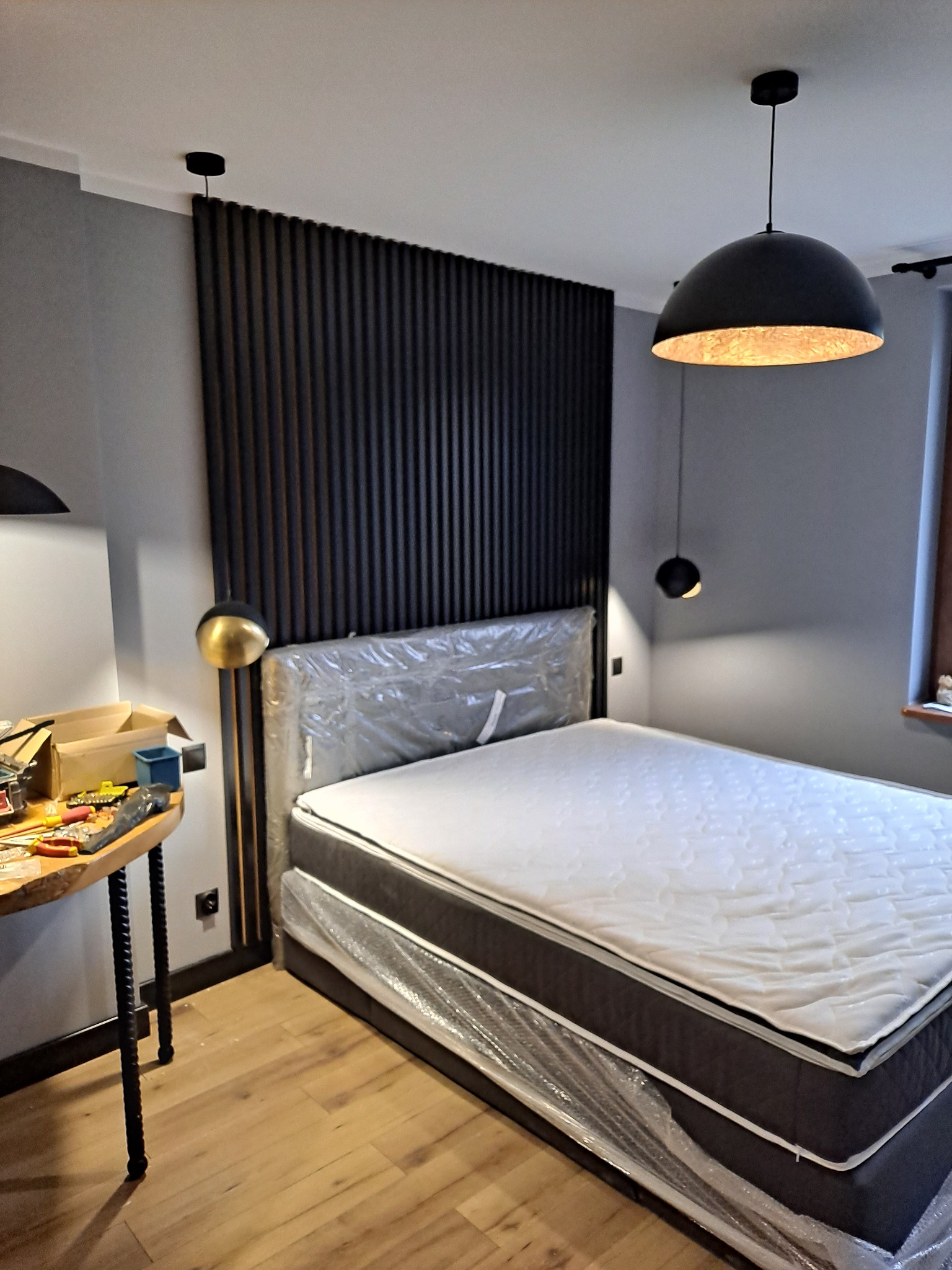 Nowe łóżko z materacem w folii ochronnej, ustawione przy ścianie z dekoracyjnymi, pionowymi panelami w kolorze ciemnego drewna. Widoczne lampy wiszące i stół roboczy z narzędziami.