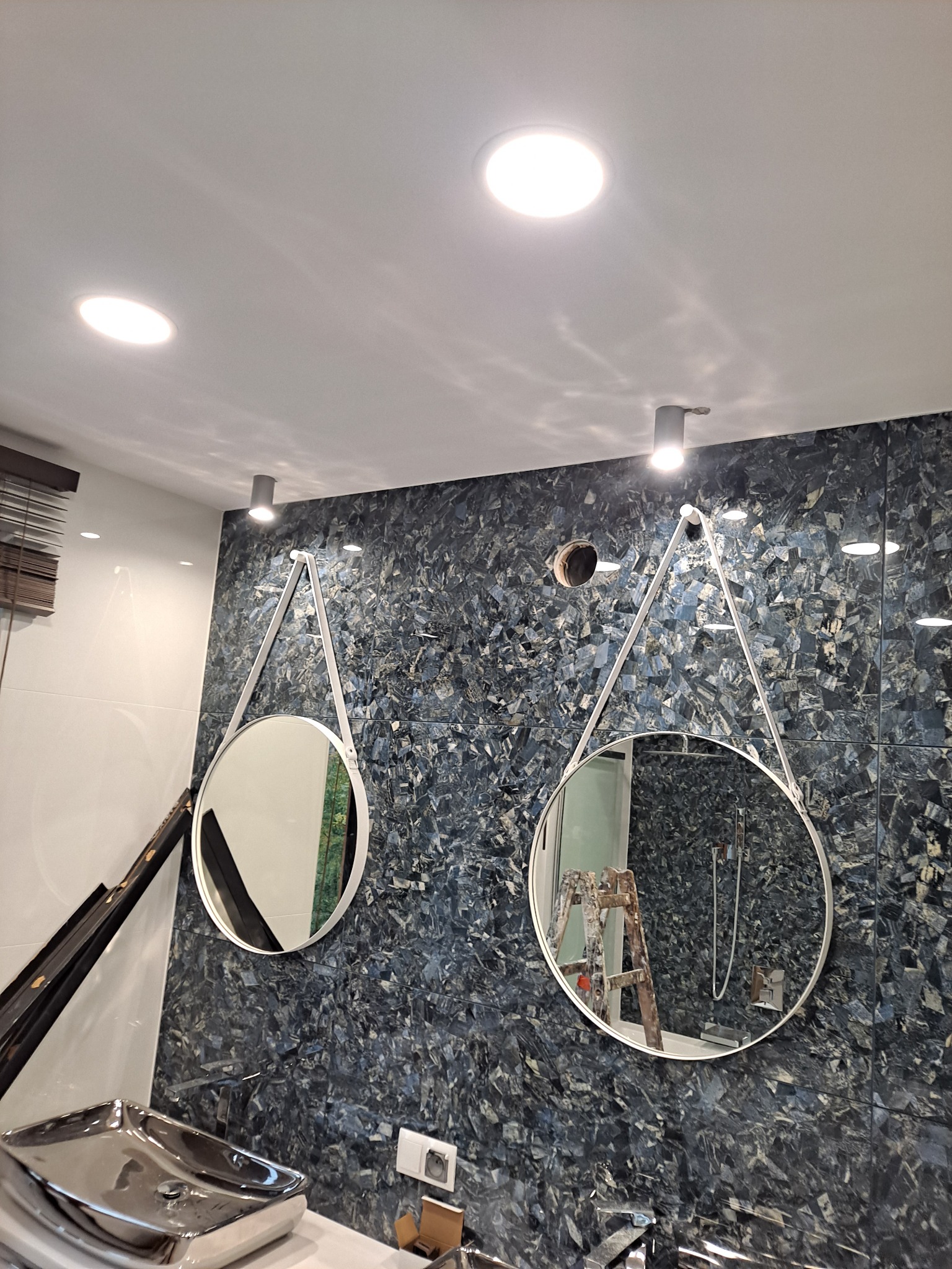 Nowoczesna łazienka z dwoma okrągłymi lustrami zawieszonymi na białych paskach, ściana wyłożona płytkami imitującymi kamień w odcieniach szarości i granatu, widoczna drabina malarska w odbiciu...