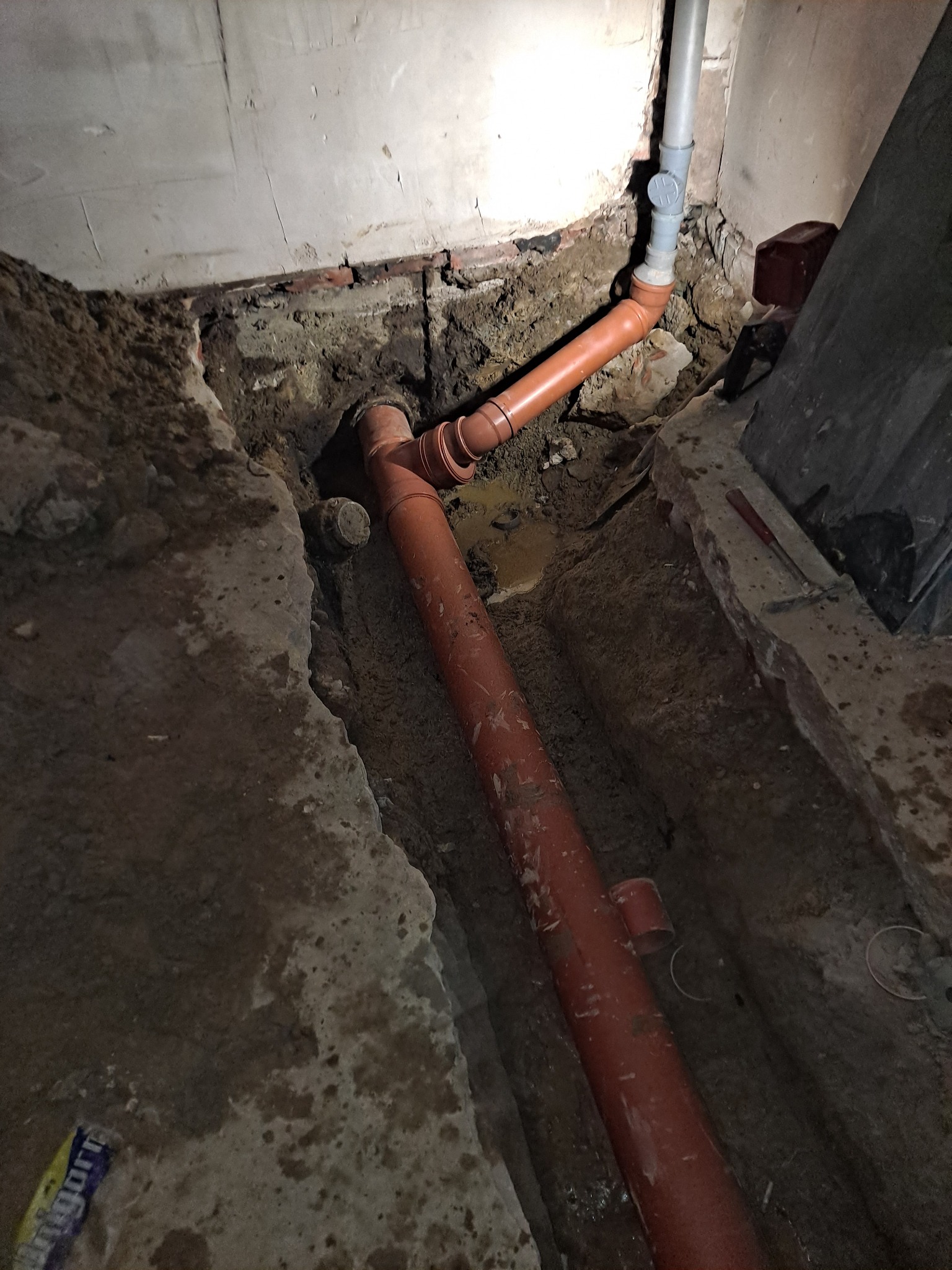 Wykonana w wykopie instalacja kanalizacyjna z rur PCV koloru ceglastego, widoczna na tle betonowych ścian i ziemi.