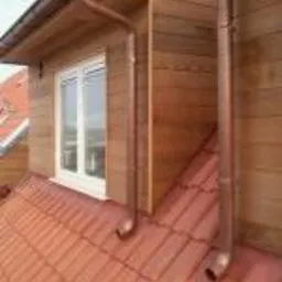 Nowe, miedziane orynnowanie budynku z dachówką ceramiczną w Gnieźnie. Widoczne okno dachowe i drewniana elewacja.