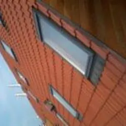 Czerwony dach z dachówki ceramicznej z oknami połaciowymi, widok z dołu na fragment budynku, błękitne niebo w tle.