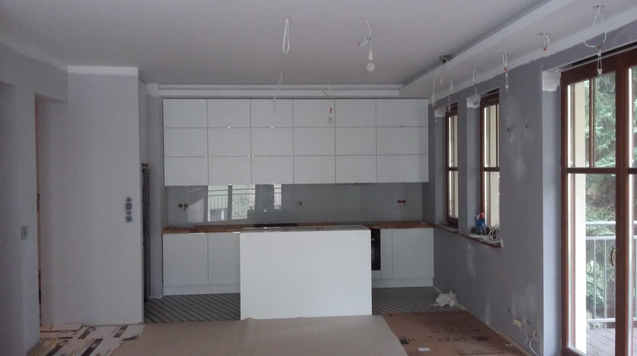 Wnętrze kuchni w trakcie prac wykończeniowych: białe szafki, wyspa, podłoga z wzorem, widoczne kable i niedokończone ściany. Okno balkonowe z widokiem na zieleń.