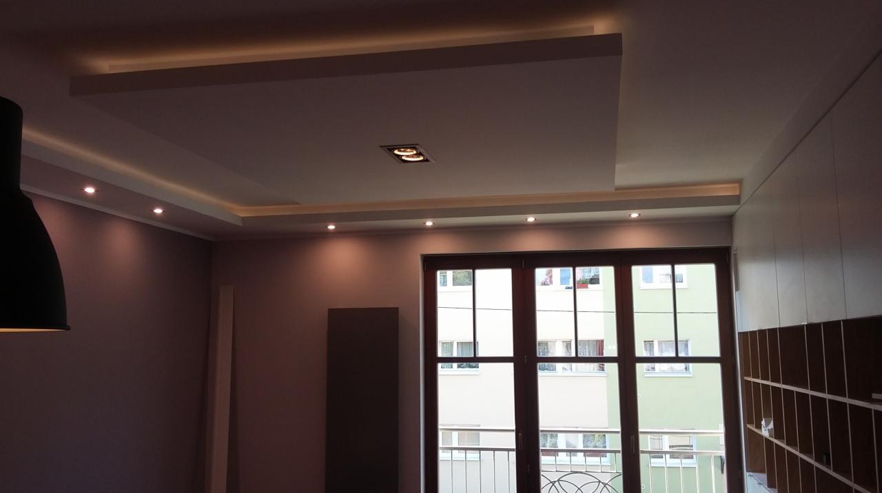 Nowoczesny sufit podwieszany z oświetleniem LED, punktowe lampy i dekoracyjne listwy. Widok na okno balkonowe i fragment szafy w zabudowie.