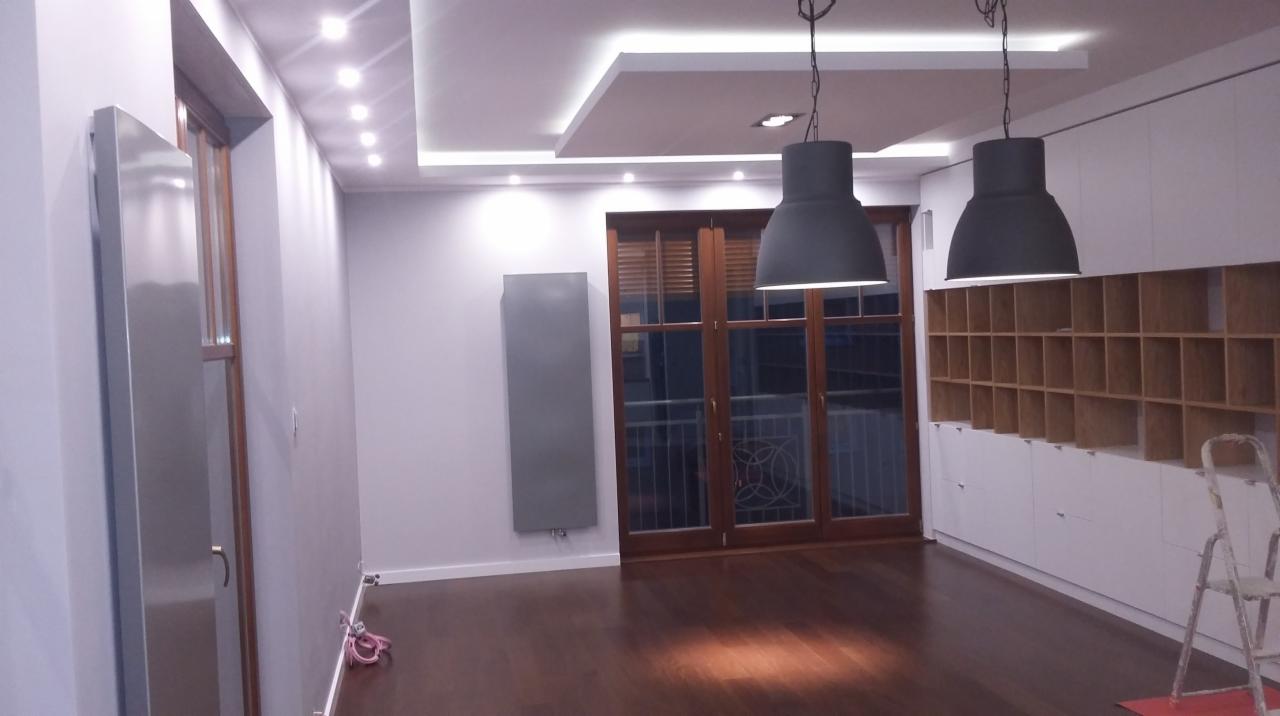 Nowoczesne wnętrze po remoncie: ciemna podłoga, białe szafki z drewnianymi półkami, szare ściany i wiszące lampy. Widoczne drzwi balkonowe i grzejnik.