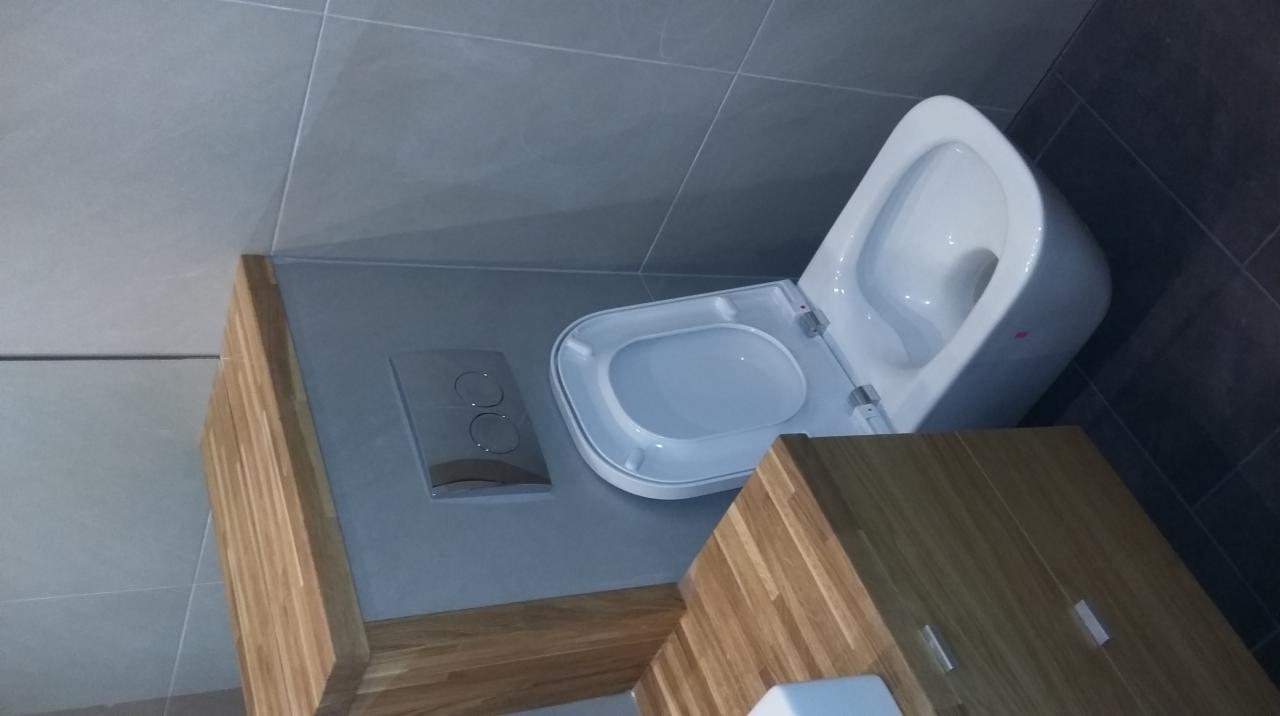 Nowoczesna toaleta podwieszana z drewnianą szafką, szarymi płytkami i chromowanym przyciskiem spłukującym. Ukończona instalacja w łazience.