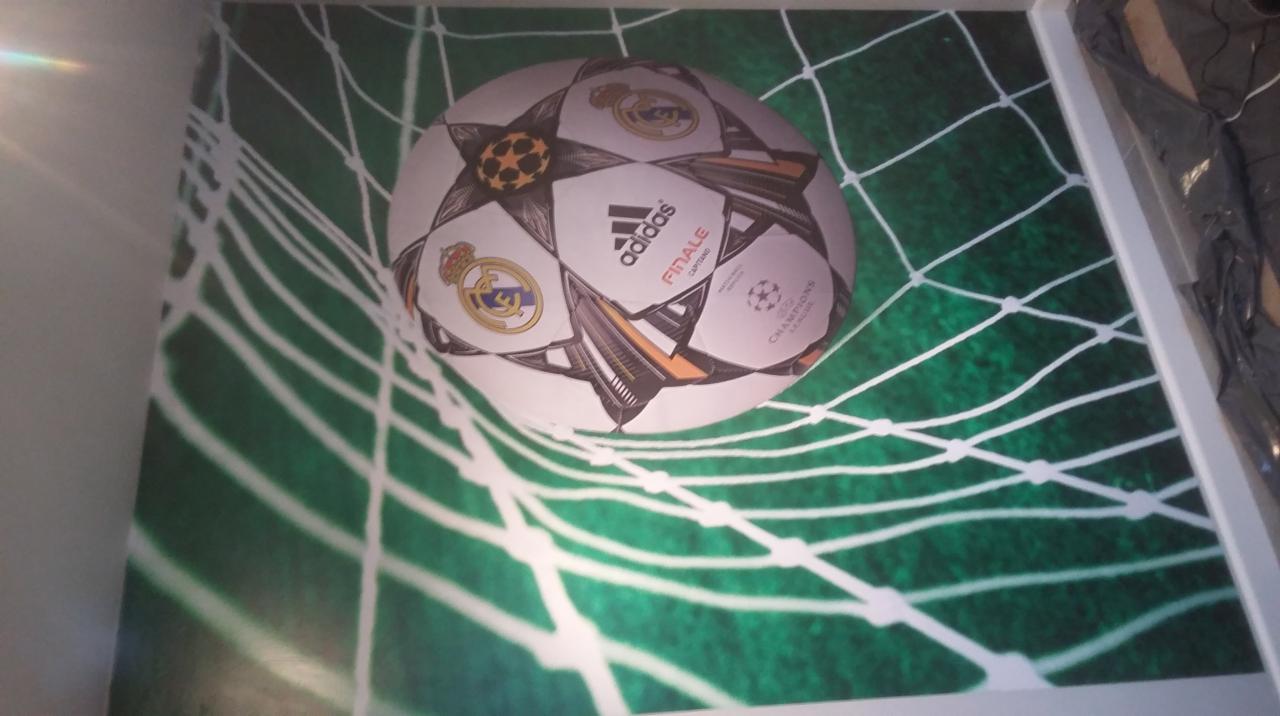 Fototapeta z motywem piłki nożnej Adidas Finale Champions League, z logiem Realu Madryt, na tle zielonej siatki, zamontowana na ścianie w pokoju.