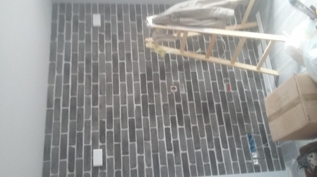 Ściana z szarej cegły dekoracyjnej, widoczne gniazdka elektryczne, drabina oparta o ścianę, kartonowe pudło. Wykończenie wnętrza w stylu industrialnym.