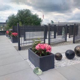 PLAST-MET Lubin Systemy Ogrodzeniowe - Nowoczesne ogrodzenie z metalowych paneli w kolorze antracytowym, oddzielające przestrzeń z betonowymi donicami z różowymi kwiatami i dekoracyjnymi kulami na białym żwirku, widoczne budynki w tle.