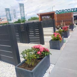 PLAST-MET Lubin Systemy Ogrodzeniowe - Nowoczesne ogrodzenie z poziomych paneli w kolorze antracytowym, zdobiące wejście do salonu sprzedaży z ekspozycją ogrodzeń; w tle widoczne flagi reklamowe producenta.