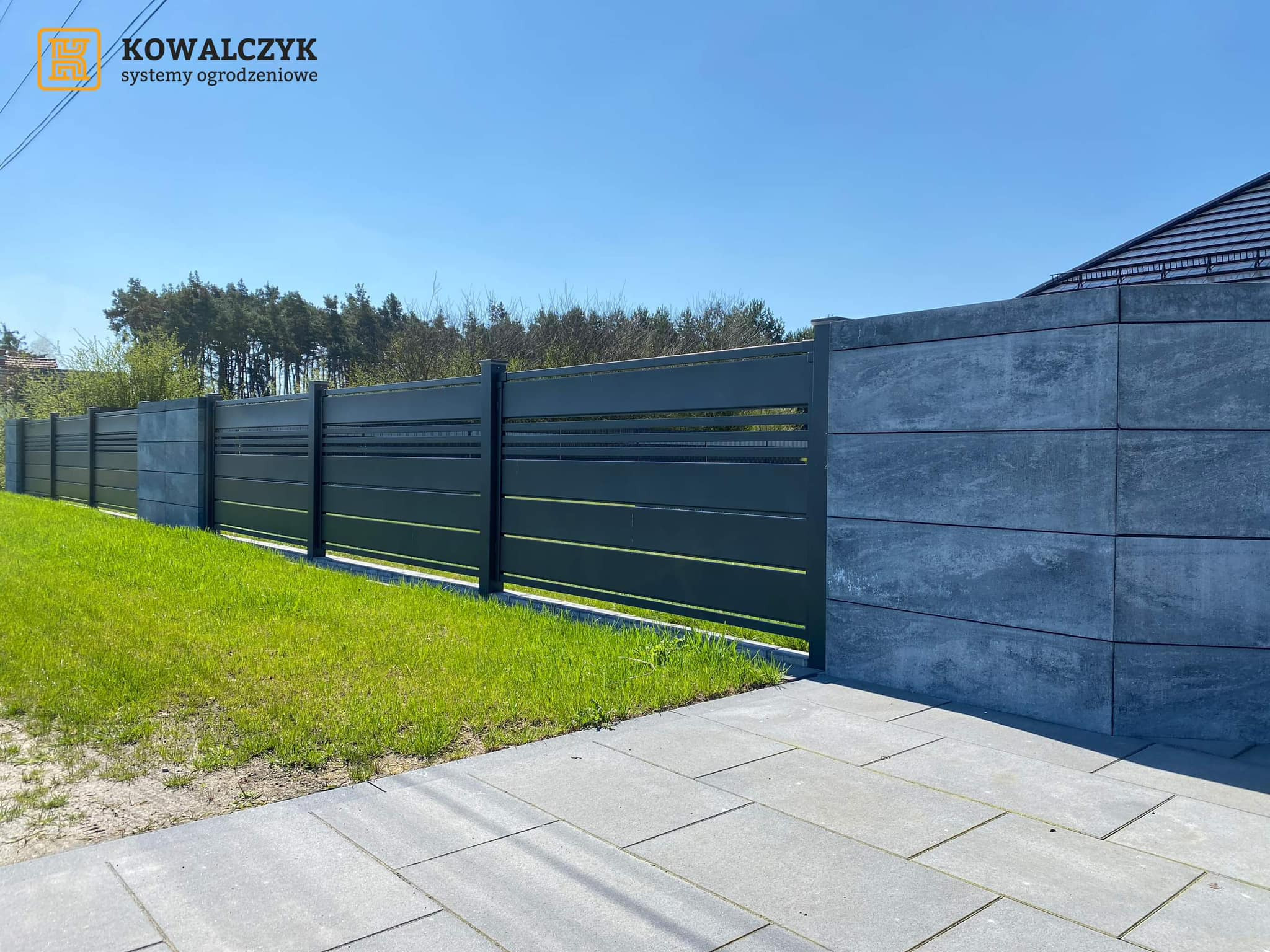 Nowoczesne ogrodzenie z poziomych, metalowych paneli w kolorze grafitowym, osadzone na betonowych słupkach, przylegające do kostki brukowej i trawnika, widok z poziomu chodnika.