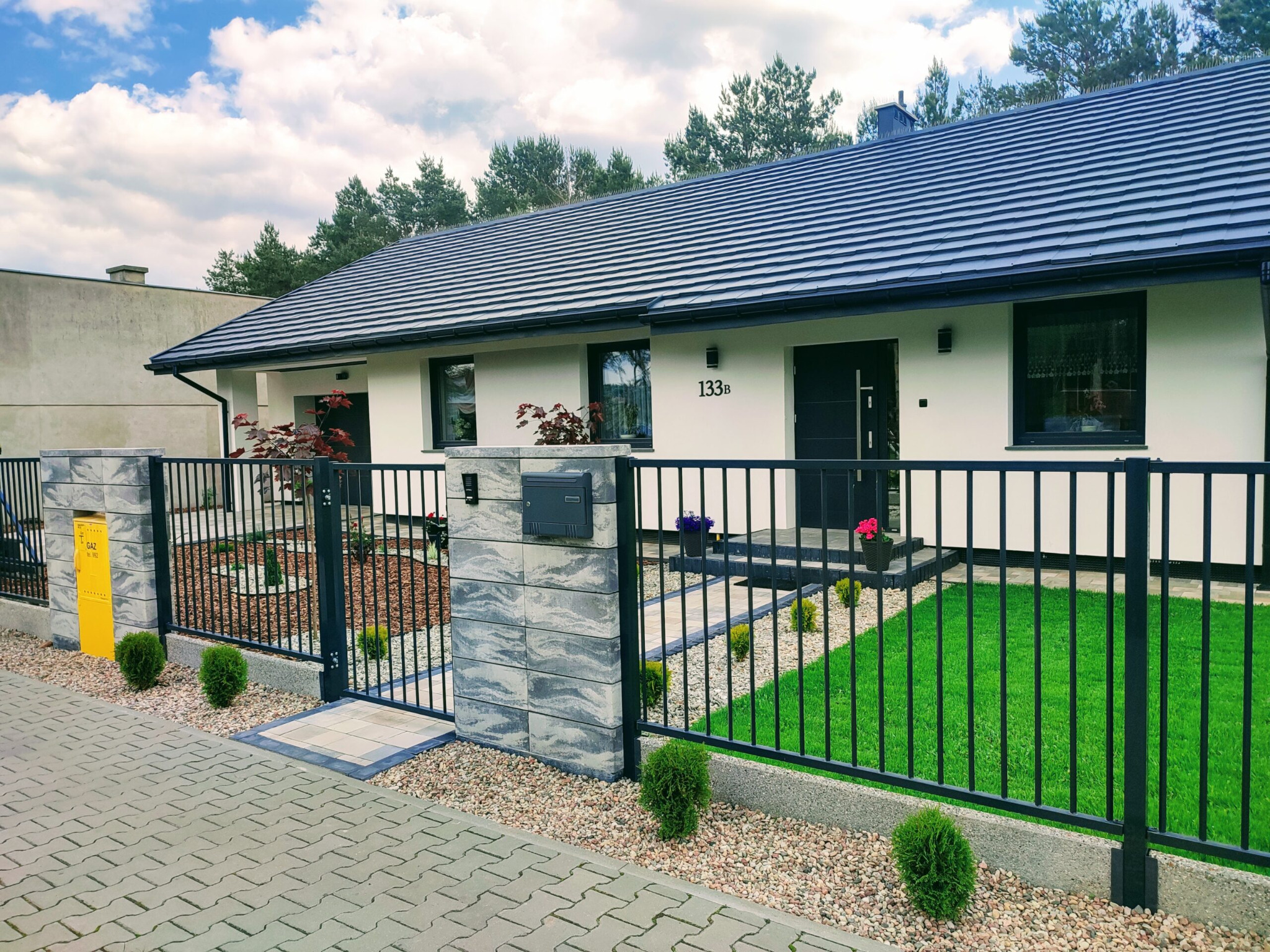 Nowoczesna posesja z czarnym ogrodzeniem sztachetowym na betonowej podmurówce, z bramką wejściową pomiędzy słupkami z szarej kostki, na tle białego domu z ciemną dachówką.