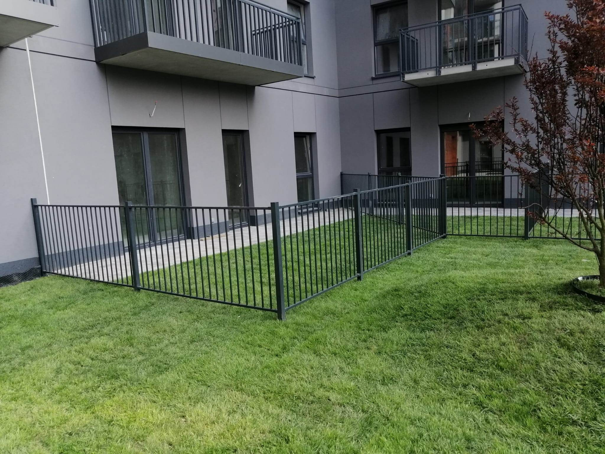 Nowoczesne, czarne ogrodzenie metalowe okalające trawnik przed budynkiem z balkonami, z widocznym drzewem z czerwonymi liśćmi po prawej stronie.