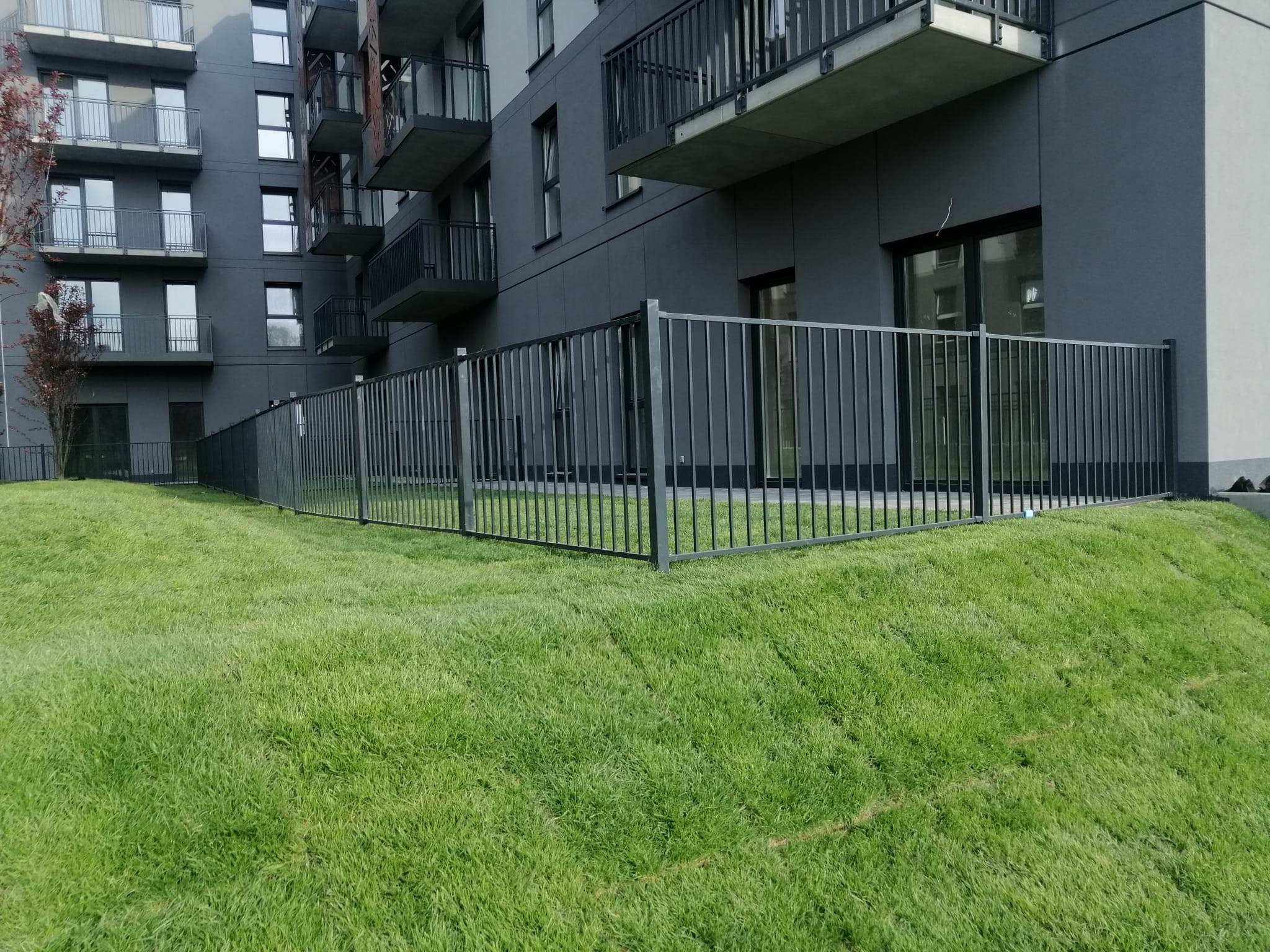 Ciemnoszare ogrodzenie metalowe z pionowych prętów okala trawnik przed nowoczesnym apartamentowcem o szarej elewacji z balkonami.