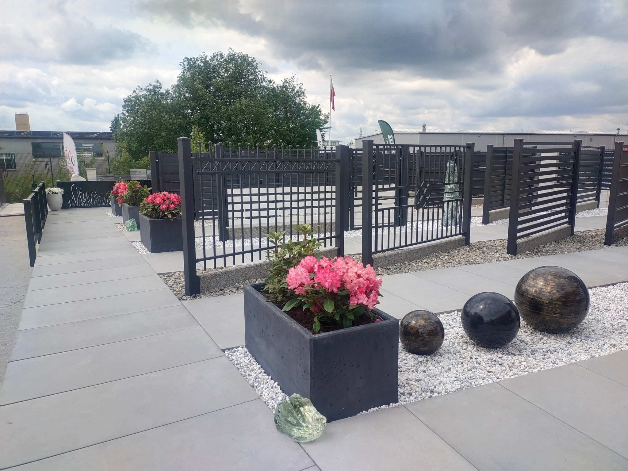 Nowoczesne ogrodzenie z metalowych paneli w kolorze antracytowym, oddzielające przestrzeń z betonowymi donicami z różowymi kwiatami i dekoracyjnymi kulami na białym żwirku, widoczne budynki w tle.