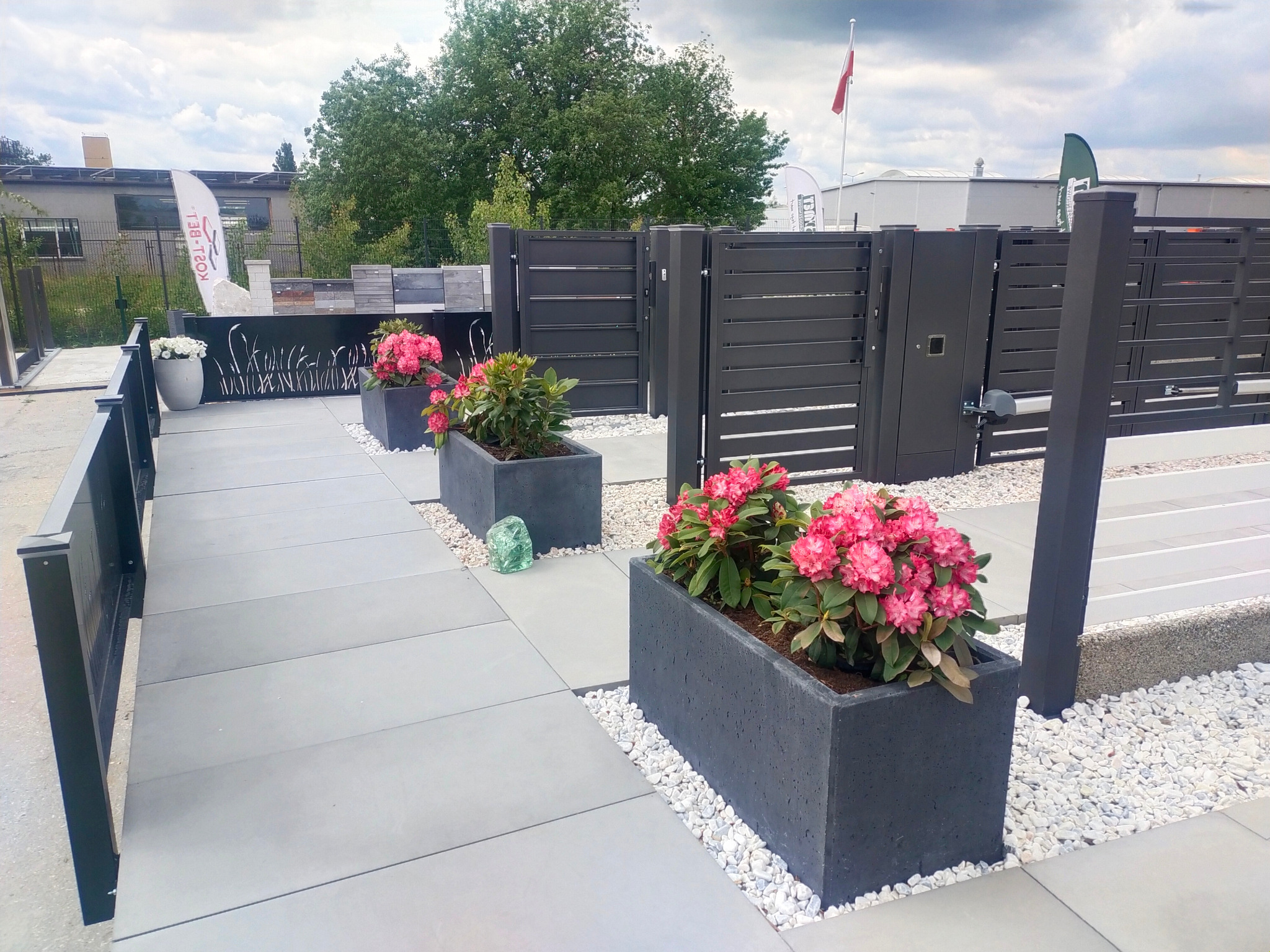 Nowoczesne ogrodzenie z poziomych paneli w kolorze grafitowym, z bramą wjazdową i furtką, ozdobione betonowymi donicami z kwitnącymi różanecznikami i białym grysem.