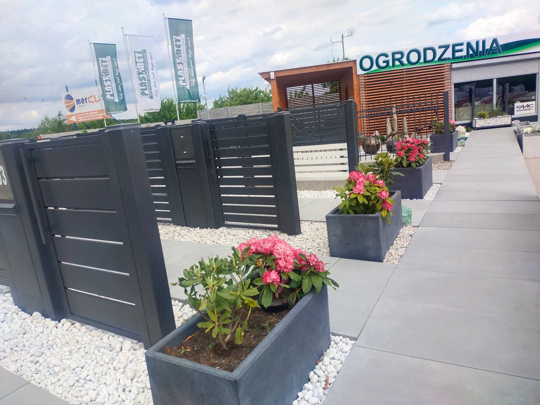 Nowoczesne ogrodzenie z poziomych paneli w kolorze antracytowym, zdobiące wejście do salonu sprzedaży z ekspozycją ogrodzeń; w tle widoczne flagi reklamowe producenta.
