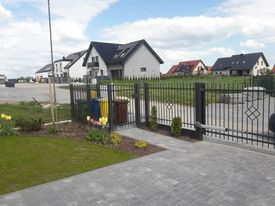 Nowoczesne ogrodzenie metalowe z bramą wjazdową na posesji z kostki brukowej, widoczne domy w tle i wiosenna zieleń.