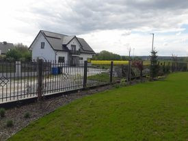 Czarne, metalowe ogrodzenie z ozdobnymi elementami w kształcie rombów, otaczające posesję z białym domem i zielonym trawnikiem w słoneczny dzień.