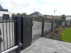 Czarna, metalowa brama wjazdowa z automatyką, widok z bliska na skrzydło bramy i słupek z domofonem, w tle ogrodzenie metalowe i dom jednorodzinny.