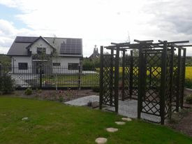 Nowoczesny dom z szarym dachem i panelami słonecznymi, otoczony czarnym, metalowym ogrodzeniem. Na pierwszym planie drewniana altana z kratownicą, ścieżka z kamieni w trawie i rabaty kwiatowe.