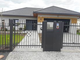 Nowoczesne ogrodzenie metalowe z bramą i furtką w kolorze antracytowym, z numerem domu 80, na tle jasnego domu z szarym dachem.