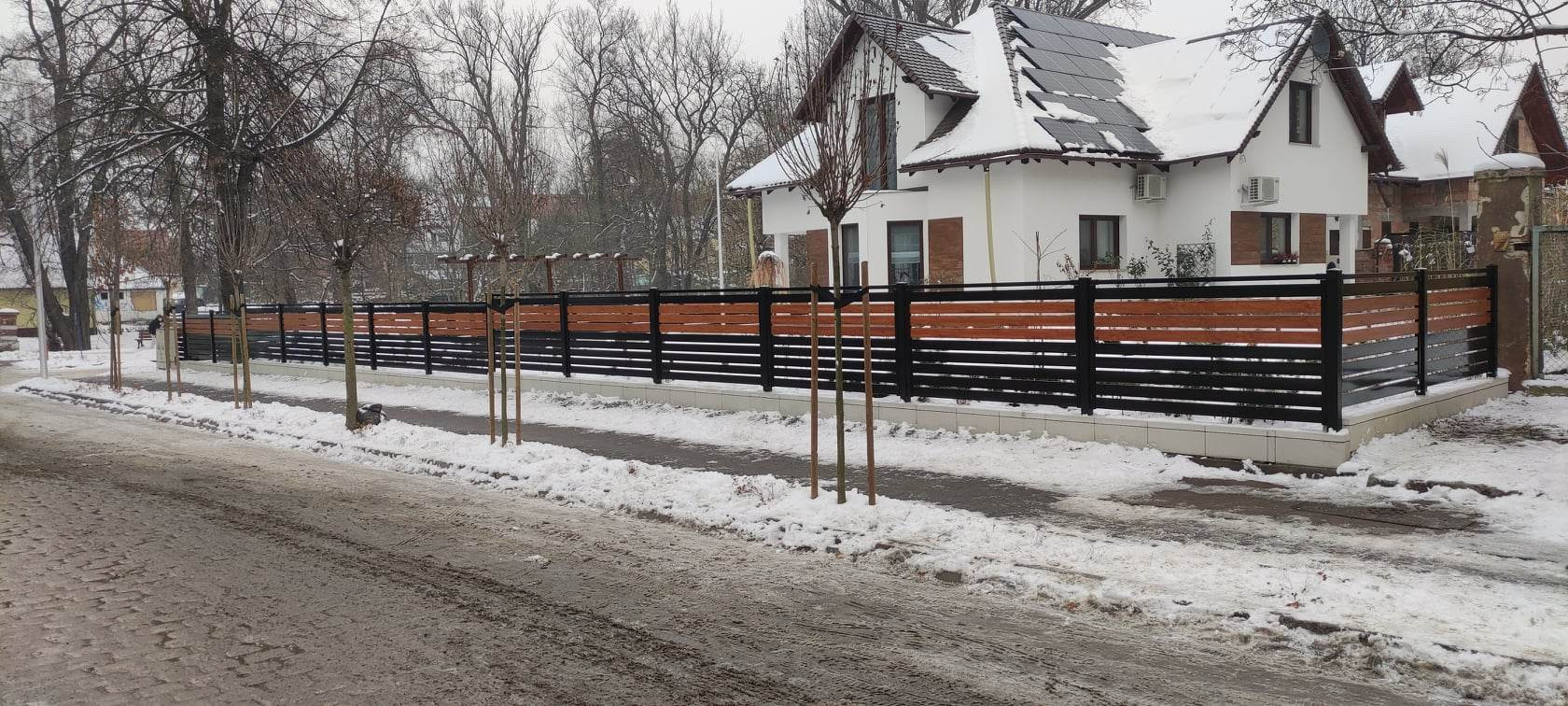 Nowoczesne ogrodzenie z poziomych desek w kolorze drewna i czarnych metalowych słupków, oddzielające posesję z białym domem z panelami słonecznymi od zasypanej śniegiem ulicy.