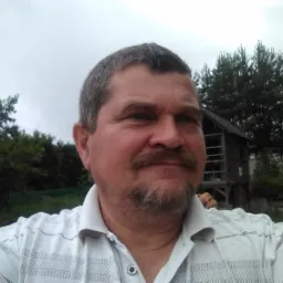 Portret mężczyzny w koszuli na tle drewnianej konstrukcji w ogrodzie, pochmurne niebo.