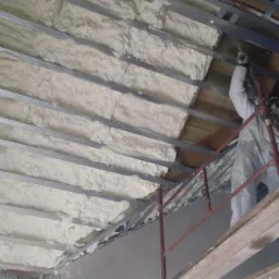 Ocieplenie poddasza pianką natryskową między metalowymi profilami. Pracownik w kombinezonie ochronnym aplikuje izolację z rusztowania.