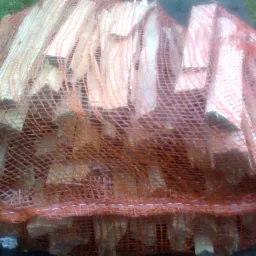 Worek z pociętym drewnem kominkowym, widoczne kawałki drewna przez pomarańczową siatkę worka, na zewnątrz, w tle trawa.